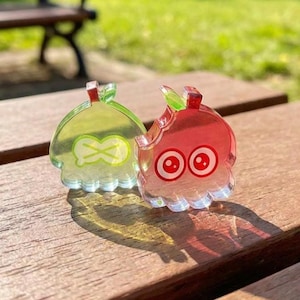 May include: Two translucent acrylic charms shaped like stylized fruit characters. One charm is lime green with a light green leaf and a white X-shaped mouth. The other charm is red with a green leaf and two large, round eyes with white highlights.