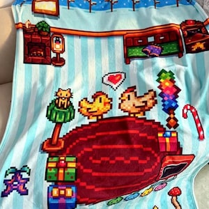 May include: A light blue throw blanket with a pixel art pattern featuring a winter scene, a red rug, two yellow chickens, a heart, gift boxes, a cat on a green post, a candy cane, a mushroom, and a star on a light blue striped background.
