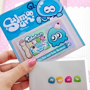 Op de afbeelding: Een Splatoon-stickervel met een blauw inkling-personage, een lang wit wezen en een dessertafbeelding met de tekst 'The One and Only Mammal'. Daaronder toont een aparte kaart vier kleine, kleurrijke, afgeronde karakterstickers.