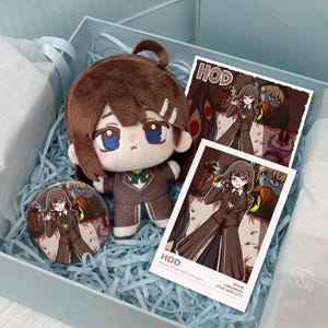 Puede incluir: Una muñeca de peluche de pelo castaño y ojos azules con traje marrón, un pin circular con un personaje de anime con traje marrón y dos impresiones cuadradas que muestran al mismo personaje con el texto HOD, BOOK, LIBRARIAN, STAR AND CITY.