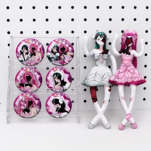 Peut inclure: Un présentoir en acrylique transparent contenant six badges ronds avec des illustrations de style anime et le texte "WORLD MY WORLD", accompagné de deux poupées en tissu avec des vêtements imprimés, l'une en robe blanche et l'autre en rose.