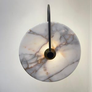 May include: A circular wall sconce made of white marble with grey veining, featuring a black metal handle. The centre is brass-coloured, with a black circular light source. The sconce is mounted on a white wall.