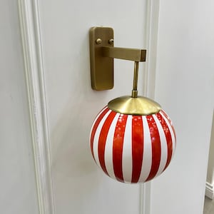 May include: A wall-mounted light fixture with a brass finish. The fixture features a round, red and white striped globe shade. The shade is suspended from a brass arm and mounted on a rectangular brass base.