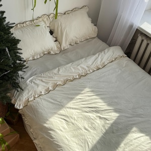 May include: A bed with cream-colored bedding, including a duvet cover and two pillowcases. The bedding has ruffled edges. A Christmas tree and wrapped gifts are visible in the corner of the room.