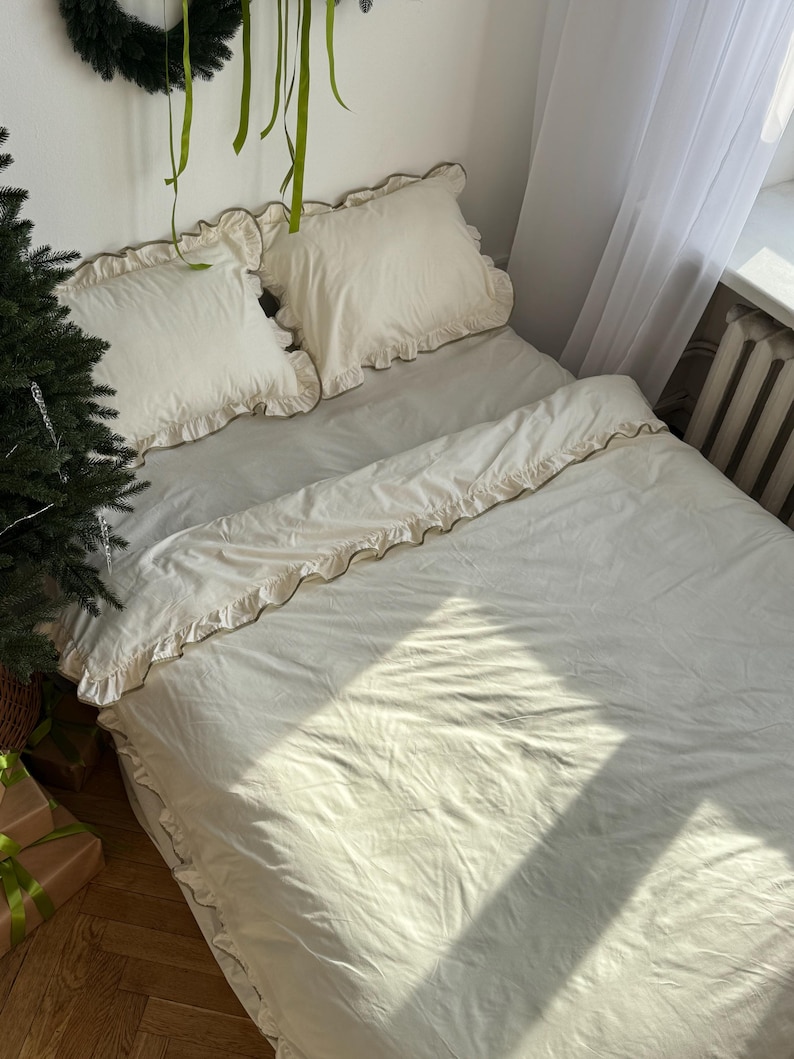 May include: A bed with cream-colored bedding, including a duvet cover and two pillowcases. The bedding has ruffled edges. A Christmas tree and wrapped gifts are visible in the corner of the room.
