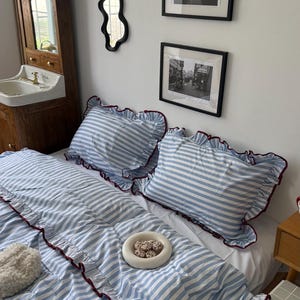 Peut inclure: Une parure de lit avec un motif à rayures bleu clair et blanc sur la housse de couette et deux taies d'oreiller assorties. Chaque pièce est dotée d'un bord à volants garni d'un tissu rouge foncé. Un petit plateau circulaire blanc est posé.