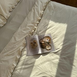 May include: Cream-colored bedding set with ruffled edges and a magazine open on top. The magazine features images of decorative objects. The bedding is neatly arranged on a bed, with sunlight casting shadows.