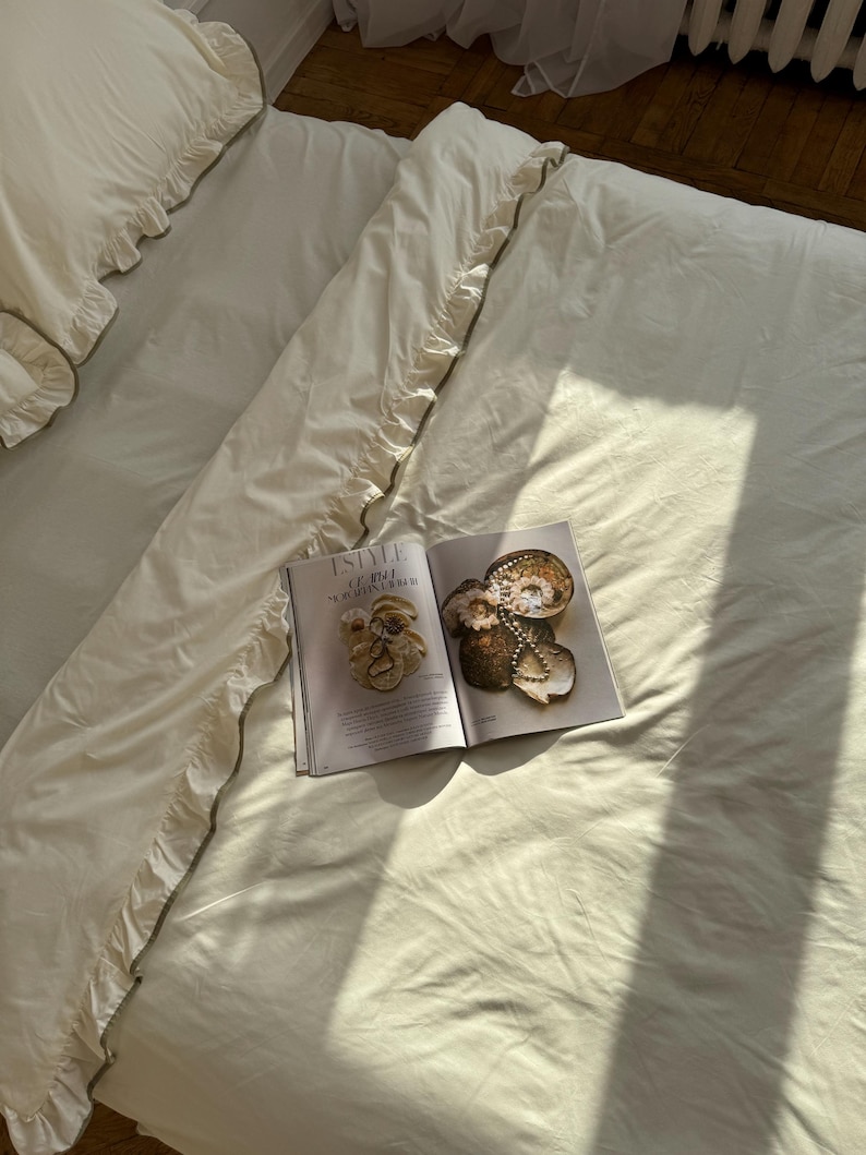 May include: Cream-colored bedding set with ruffled edges and a magazine open on top. The magazine features images of decorative objects. The bedding is neatly arranged on a bed, with sunlight casting shadows.