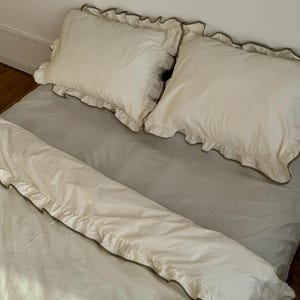 May include: A bed with a light gray sheet and two white pillows with ruffled edges. A white duvet cover with ruffled edges is also on the bed. The ruffles are trimmed with a darker color.