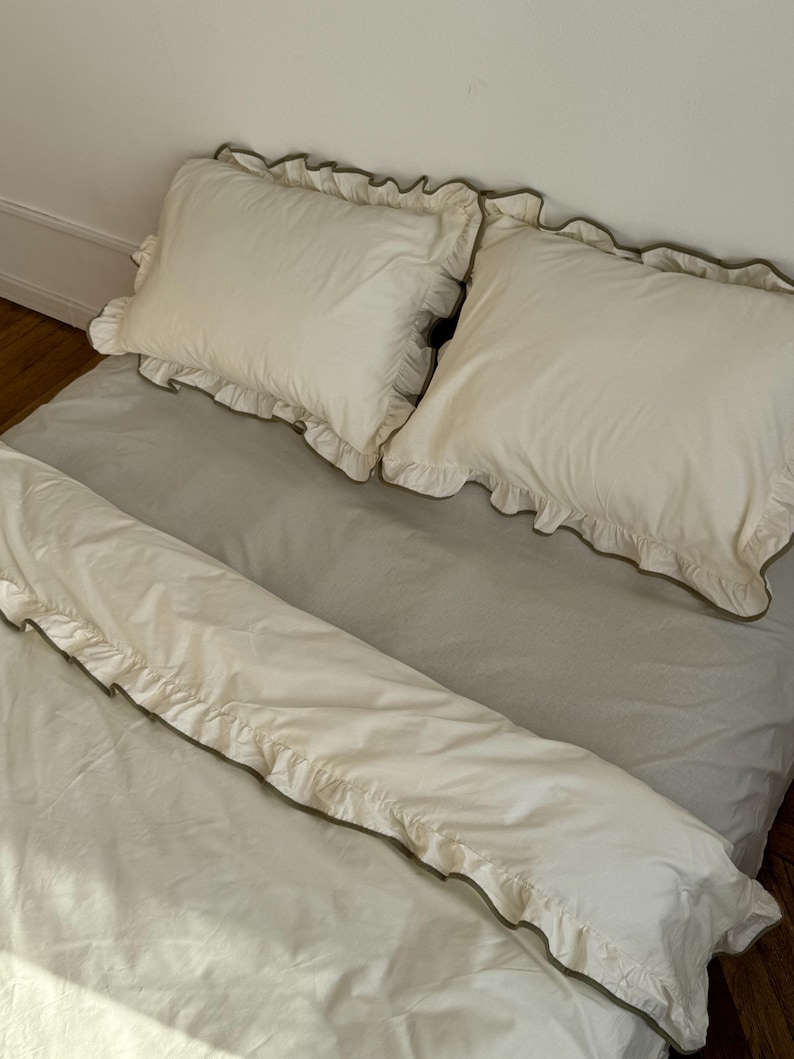 May include: A bed with a light gray sheet and two white pillows with ruffled edges. A white duvet cover with ruffled edges is also on the bed. The ruffles are trimmed with a darker color.