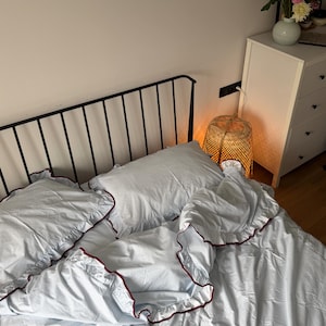 May include: Bedding set with a light blue and white pattern, trimmed with a dark red ruffle. The bed has a black metal headboard. A white chest of drawers with a vase of flowers is visible.