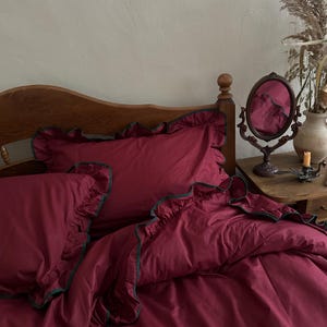 Puede incluir: Juego de cama burdeos con bordes con volantes y ribete verde oscuro. El juego incluye una funda nórdica y fundas de almohada, dispuestas sobre una estructura de cama de madera. Un espejo decorativo y un jarrón son visibles en el fondo.