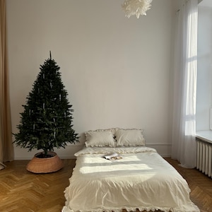 May include: A bedroom scene featuring a bed with cream-colored bedding and ruffled edges. Two pillows are on the bed, and a magazine rests on the duvet. A Christmas tree stands in the corner, and the room has hardwood floors.
