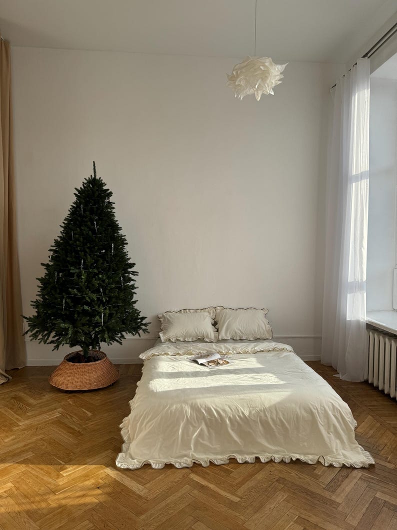 May include: A bedroom scene featuring a bed with cream-colored bedding and ruffled edges. Two pillows are on the bed, and a magazine rests on the duvet. A Christmas tree stands in the corner, and the room has hardwood floors.