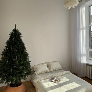May include: A bedroom scene featuring a Christmas tree in a woven basket, a bed with cream-colored bedding, and a hanging light fixture. The bed has two pillows and a book. Sunlight streams through a sheer curtained window, illuminating the room.