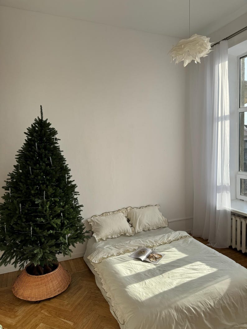 May include: A bedroom scene featuring a Christmas tree in a woven basket, a bed with cream-colored bedding, and a hanging light fixture. The bed has two pillows and a book. Sunlight streams through a sheer curtained window, illuminating the room.