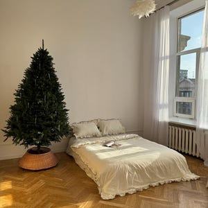May include: A bedroom scene featuring a bed with cream-colored bedding and ruffled edges. A green Christmas tree stands to the left, with a woven basket base. The room has a large window with sheer curtains and a herringbone wood floor.