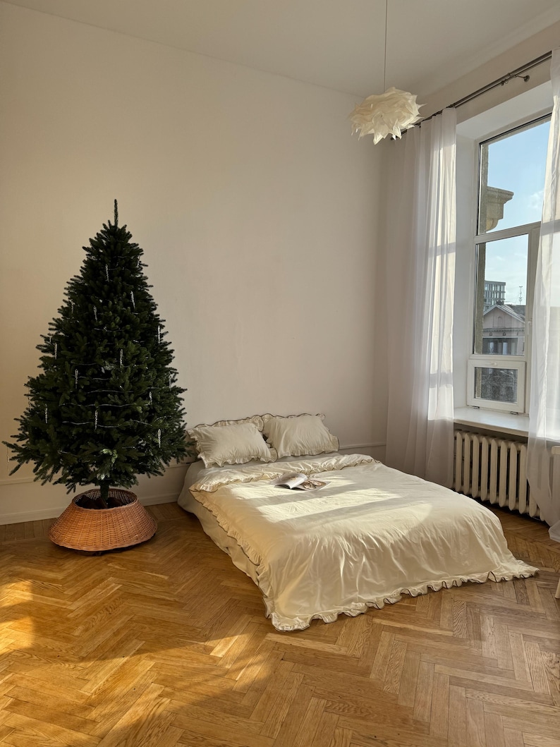 May include: A bedroom scene featuring a bed with cream-colored bedding and ruffled edges. A green Christmas tree stands to the left, with a woven basket base. The room has a large window with sheer curtains and a herringbone wood floor.