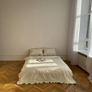 May include: A cream-colored bed with ruffled bedding and two matching pillows. The bed is in a room with a wooden floor and a large window with white curtains. A white paper-like light fixture hangs from the ceiling.