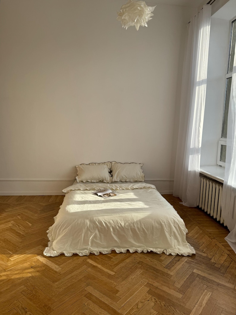 May include: A cream-colored bed with ruffled bedding and two matching pillows. The bed is in a room with a wooden floor and a large window with white curtains. A white paper-like light fixture hangs from the ceiling.