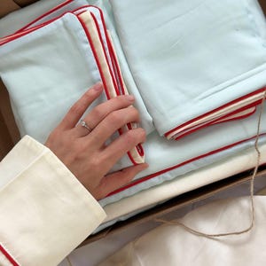 May include: A set of light blue bedding with red trim, neatly folded inside a cardboard box. The bedding includes pillowcases and sheets. A hand wearing a ring is touching the bedding. The sleeve of a cream-colored shirt with red trim is visible.