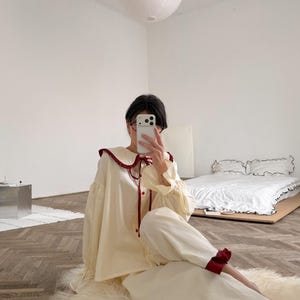 以下が含まれることがあります： クリーム色のパジャマセット。大きな丸襟が付いた長袖のボタンダウンシャツと、お揃いのパンツで構成されています。襟、袖口、パンツの裾には、ダークレッドのフリルがあしらわれています。