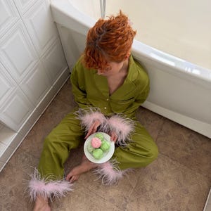 以下が含まれることがあります： オリーブグリーンのボタンダウン長袖シャツとパンツのツーピースパジャマセット。袖口とパンツの裾には、柔らかいライトピンクの羽毛があしらわれています。小さな白い皿には、パステルカラーのスイーツが3つ載っています。
