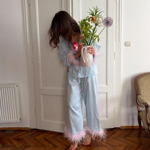 May include: A light blue pyjama set consisting of a button-down long-sleeve shirt and matching trousers, both featuring light pink feather trim at the cuffs and hems. The shirt has a collar and the trousers have a relaxed, straight-leg fit.