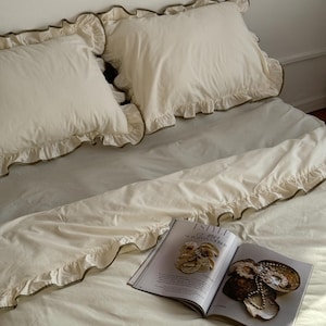 May include: Cream-colored bedding set with ruffled edges and a magazine. The set includes two pillowcases and a duvet cover, all with a delicate ruffle trim. The magazine is open, displaying images of jewelry and accessories.