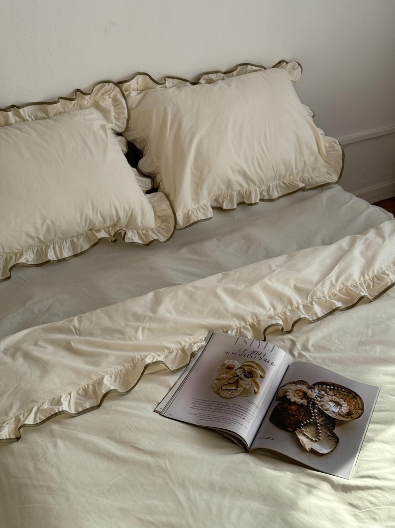 May include: Cream-colored bedding set with ruffled edges and a magazine. The set includes two pillowcases and a duvet cover, all with a delicate ruffle trim. The magazine is open, displaying images of jewelry and accessories.