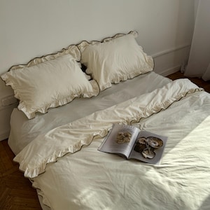 May include: A bed with cream-colored bedding, including two ruffled pillowcases and a duvet cover. The bedding has a subtle green undertone. An open magazine rests on the duvet. The room has a minimalist aesthetic with a neutral color palette.