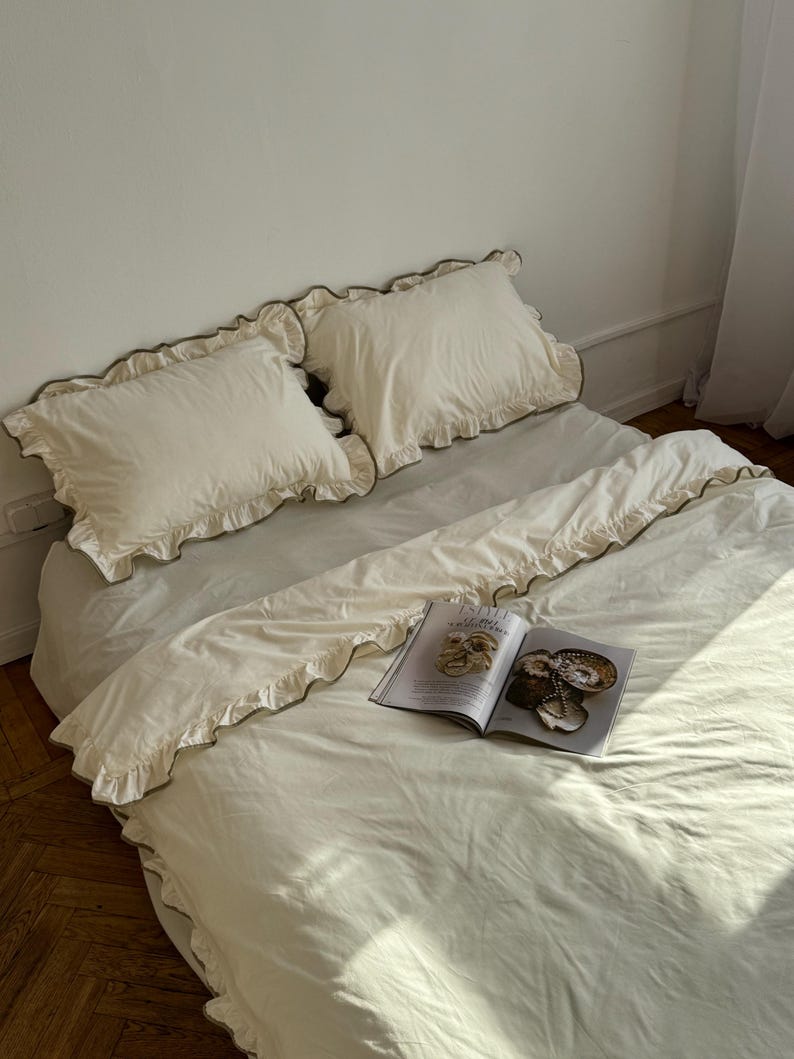 May include: A bed with cream-colored bedding, including two ruffled pillowcases and a duvet cover. The bedding has a subtle green undertone. An open magazine rests on the duvet. The room has a minimalist aesthetic with a neutral color palette.