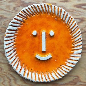 Peut inclure: Un soleil rond en céramique orange avec un visage souriant. Le soleil a une bordure blanche avec des lignes rayonnantes. Le visage comporte deux points blancs pour les yeux, une ligne verticale blanche pour le nez et une ligne blanche incurvée pour la bouche.