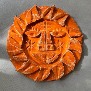 May include: A circular orange ceramic wall plaque featuring a stylised sun face. The design includes embossed almond-shaped eyes, a vertical nose, a curved mouth, and radiating triangular rays around the perimeter with a glossy finish.