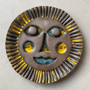 May include: A ceramic sun-shaped wall hanging with a smiling face. The sun has a brown center with blue eyes and a blue mouth. Yellow accents highlight the sun's rays and cheeks. The sun's diameter is approximately 20 cm.