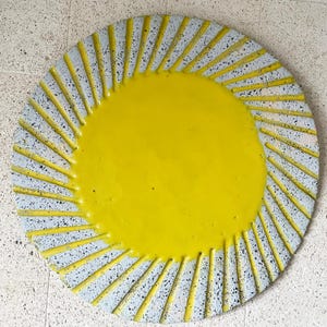 May include: A circular decorative object featuring a solid, bright yellow centre surrounded by a speckled light grey border. The border is textured with raised yellow lines radiating outwards from the centre towards the outer edge of the circle.