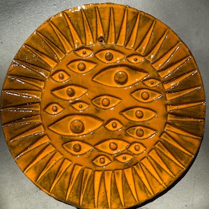 May include: A circular ceramic wall plaque glazed in a vibrant mustard yellow with a glossy finish. The surface features a relief design of a sun with radiating triangular rays and numerous embossed eyes scattered across the centre.