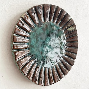 May include: Oval ceramic decorative plate with a textured, ridged rim in a dark brown glaze. The centre of the plate has a mottled teal and green glaze, creating a unique, handcrafted appearance. The plate is a decorative item.