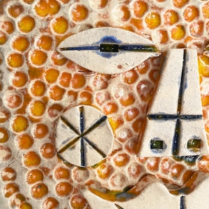 May include: Close-up of a textured ceramic artwork with a face-like design. The background features orange and yellow bubble-like shapes. The face has white, blue, and green accents, with an eye, a nose, and a mouth.