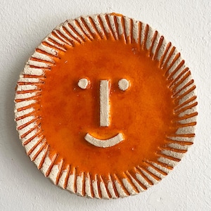 May include: A circular, orange ceramic sun with a smiling face. The sun has a textured, off-white border with radiating lines. The face features two white dots for eyes, a vertical line for a nose, and a curved line for a mouth.