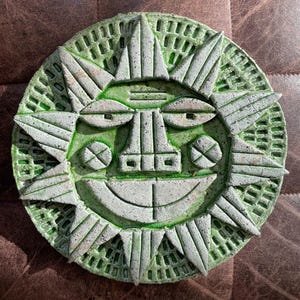 Könnte beinhalten: Eine kreisförmige Wandplatte aus Keramik mit einem stilisierten Sonnengesicht in einer texturierten, gesprenkelten hellgrauen Oberfläche. Der Hintergrund ist eine leuchtend grüne Glasur mit Gittermuster. Die Sonne hat dreieckige Strahlen.