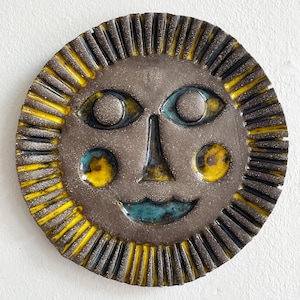 Könnte beinhalten: Keramik-Wandbehang in Sonnenform mit lächelndem Gesicht. Die Sonne hat eine braune Mitte mit blauen Augen und einem blauen Mund. Gelbe und schwarze Strahlen umgeben das Gesicht. Die Sonne hat eine strukturierte Oberfläche.