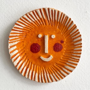 May include: A circular, orange ceramic sun with white rays extending outwards. The sun features a white nose and smile, two white dots for eyes, and two red cheek spots. This is a decorative item.