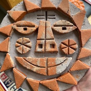 Könnte beinhalten: Eine kreisförmige graue Betonplatte mit einem Sonnengesicht-Design aus eingelegten Terrakotta-Tonstücken. Das Gesicht besteht aus dreieckigen Strahlen, mandelförmigen Augen, kreisförmigen Wangen und einem breiten, geschwungenen Mund.