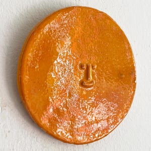 Puede incluir: Una pieza de cerámica redonda y naranja con un acabado brillante. La pieza presenta un diseño de cara simple con una forma de 'T' para los ojos y una línea curva para la boca. La cerámica tiene una superficie texturizada y está montada en una pared blanca.