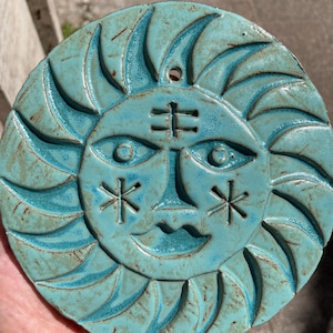 Könnte beinhalten: Eine kreisförmige, türkisfarbene Keramik-Wandplatte mit einem stilisierten Sonnengesicht und geschnitzten Details. Das Design umfasst ein zentrales Stirnsymbol, mandelförmige Augen, sternförmige Wangenmarkierungen und geschwungene Strahlen.