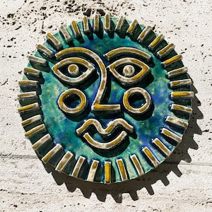 Puede incluir: Un colgante de pared con forma de cara de sol de cerámica. La cara es de color verde azulado y azul con detalles en amarillo y blanco. El sol tiene una boca sonriente, ojos redondos y una nariz. Los rayos del sol son rectangulares.