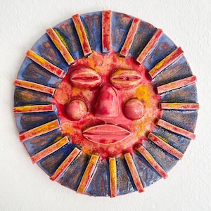 May include: A ceramic sun face wall hanging with a red, yellow, and blue colour scheme. The sun has a red face with a nose, eyes, and mouth. The sun's rays are rectangular and alternate between red and yellow. The background is blue.
