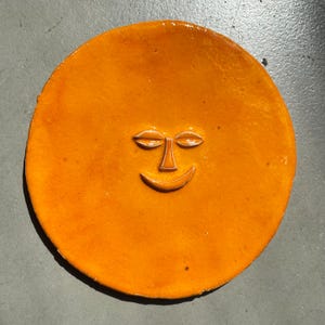 Puede incluir: Un plato redondo de cerámica naranja con un diseño de cara sonriente. La cara presenta ojos cerrados y una boca curvada, todo en un tono naranja más claro. El plato es de color sólido con un acabado brillante, y probablemente está hecho a mano.