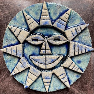 May include: A circular ceramic wall plaque featuring a stylised sun face with a textured, light blue and cream-coloured glaze. The sun has triangular rays extending outwards, almond-shaped eyes, and a wide, smiling mouth with dark blue accents.